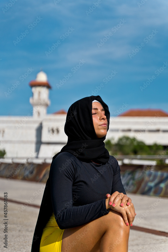 Portrait of muslim woman wearing a hijab praying isolated with mosque background. Vertical view of arabic woman outdoors. Muslim women, religion and equality concept.