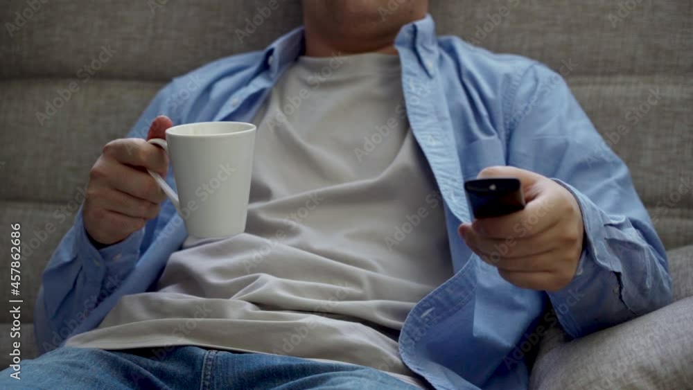 A man 30-40 years old sits on the sofa and turns on the TV from the remote control A man 30-40 years old sits on the sofa and turns on the TV from the remote control