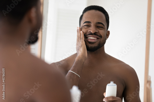 African shirtless man do morning self-hygiene routine holds bottle apply skincare cosmetics product, after shave moisturizing lotion, enjoy smooth and flawless facial skin. Skincare, grooming concept photo