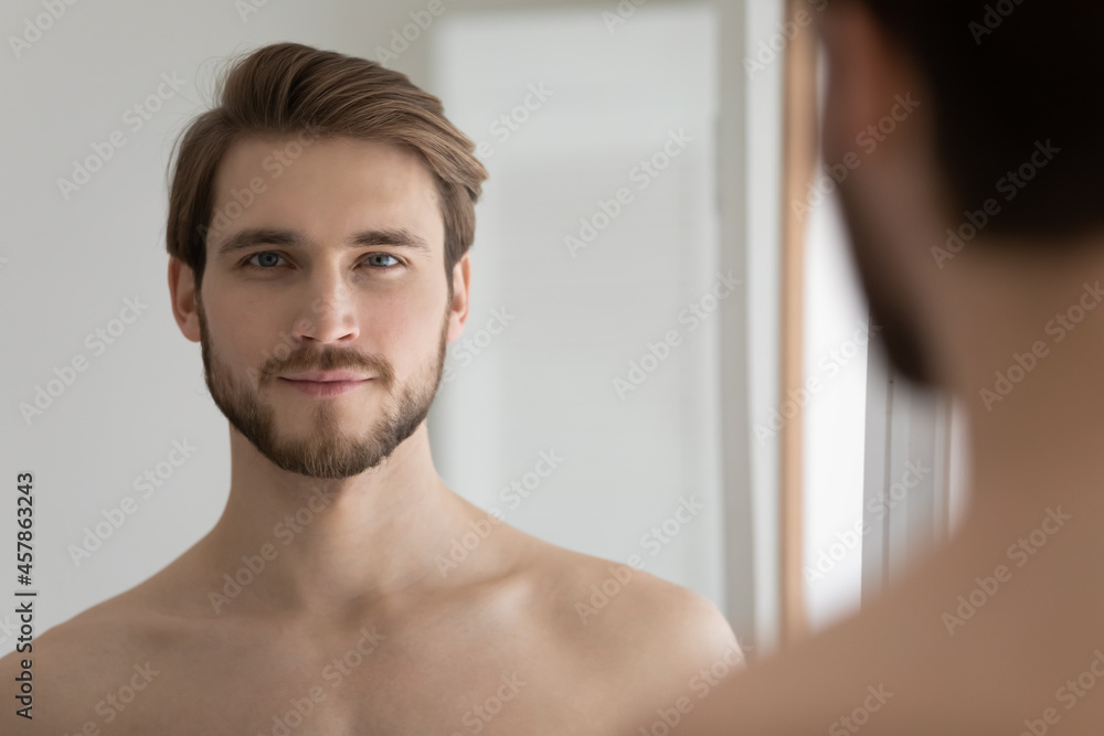 Fototapeta premium Head shot portrait young handsome good-looking groomed shirtless serious man standing in front of mirror looking at camera feels confident. Male cosmetics and beauty, morning routine, skincare concept