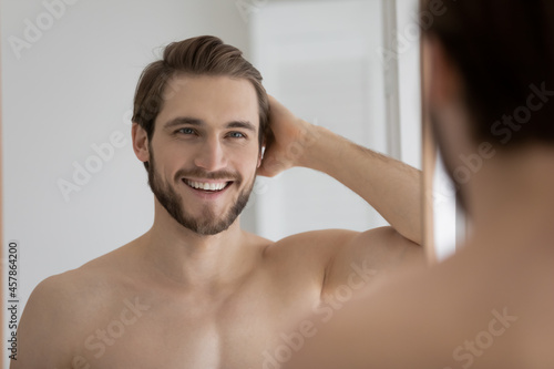 Handsome shirtless man admires his reflection in mirror, feels satisfied with quality of skin, shiny hair after morning routine of personal grooming. Cosmetics for men, skincare and haircare concept