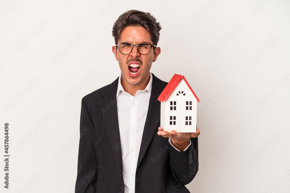 Young mixed race business man holding a toy house isolated on white background screaming very angry and aggressive. Young mixed race business man holding a toy house isolated on white background screaming very angry and aggressive.