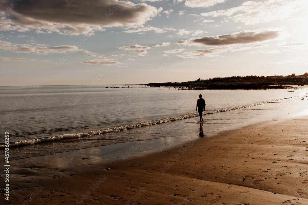 Naklejka premium Sunset on a beach in Ireland. A person is walking on the beach of Silverstrand near Galway. Golden hour beach image with clouds and a silhouette of a man. 