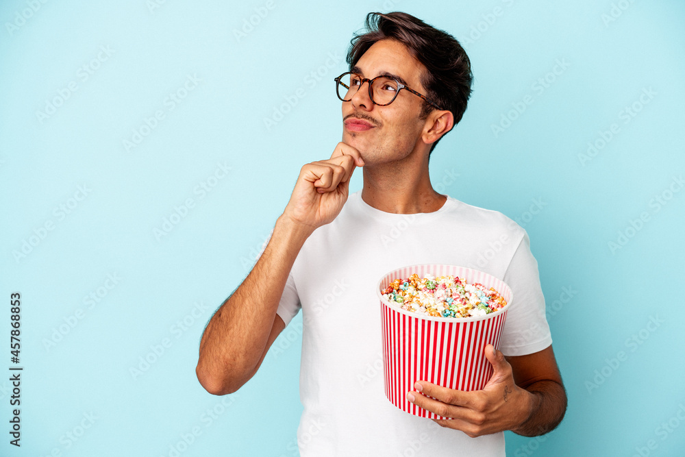 Young mixed race man eating popcorns isolated on blue background looking sideways with doubtful and skeptical expression.