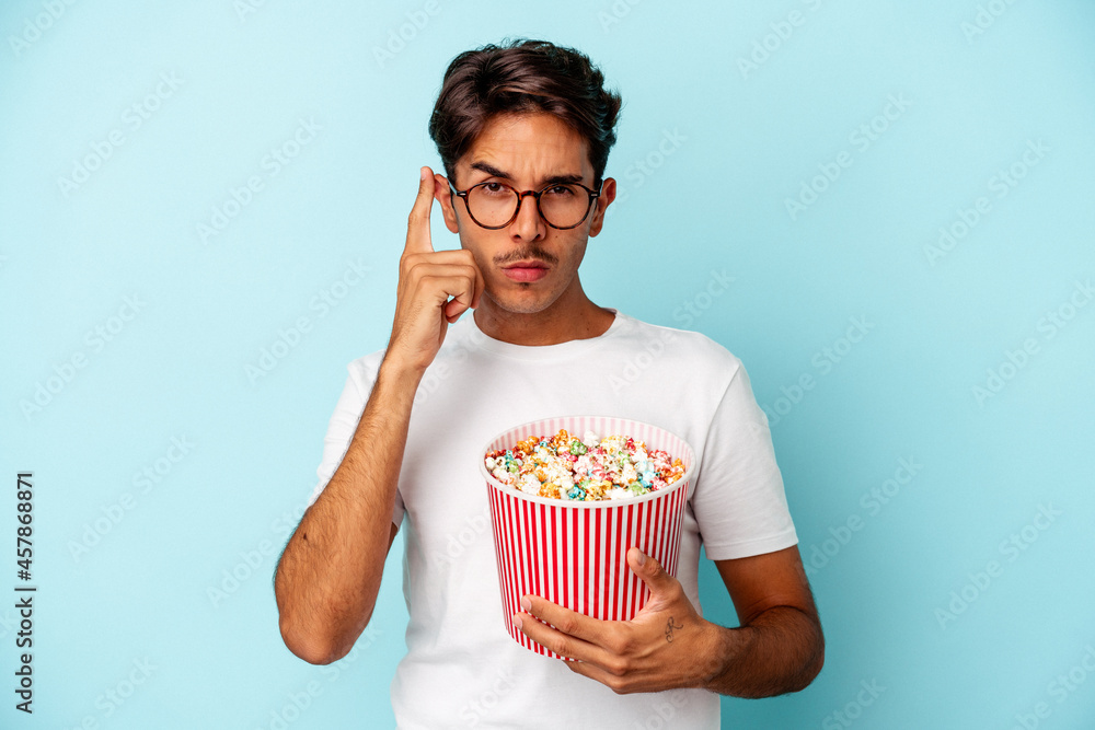 Young mixed race man eating popcorns isolated on blue background pointing temple with finger, thinking, focused on a task.
