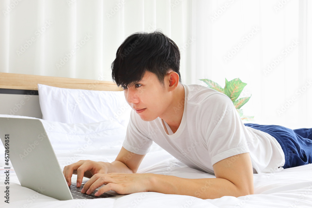 Young freelance Asian man working from home using laptop computer while sitting on the bed with copy space Young freelance Asian man working from home using laptop computer while sitting on the bed with copy space
