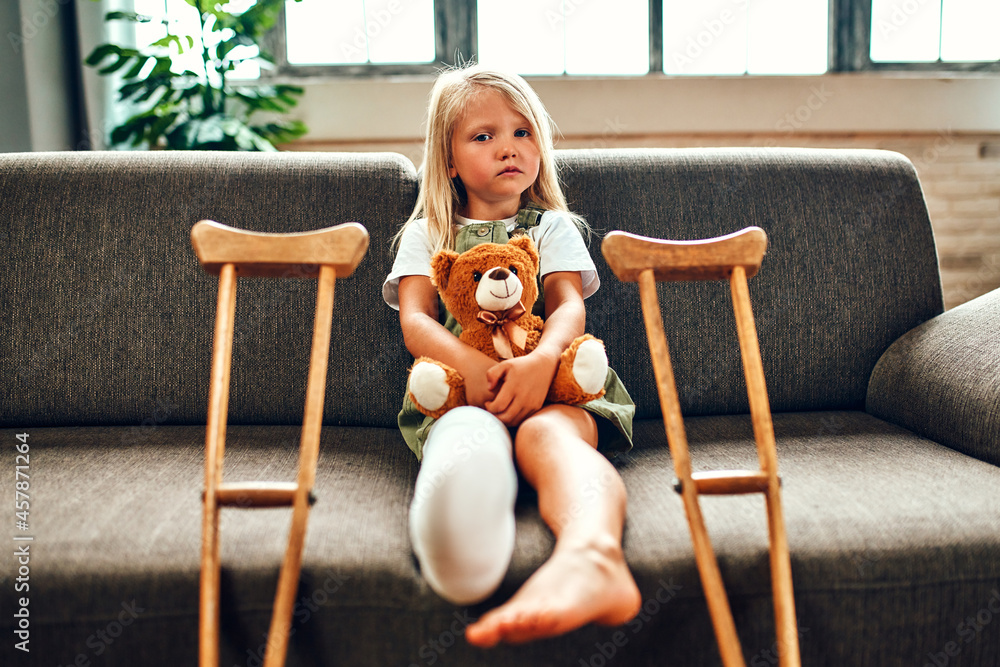 A cute sad little girl with a broken leg in a cast, sits on the couch