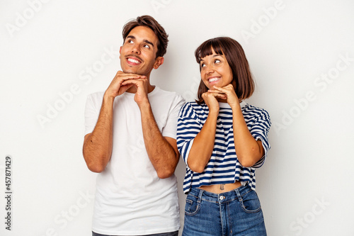 Young mixed race couple isolated on white background keeps hands under chin, is looking happily aside.
