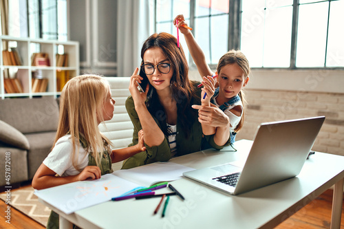 Obraz na plátně A business woman and a mother are trying to work on a laptop when her little daughters are playing, fooling around and interfering with her