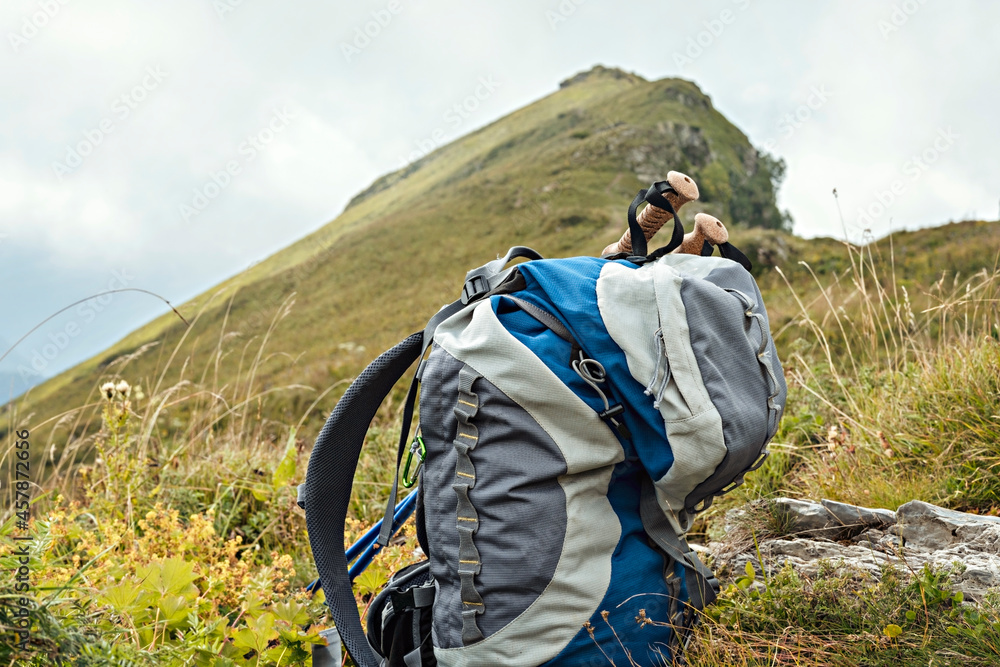 big gray blue hiking backpack, trekking poles among green plants on the background of a mountain peak, hike active healthy lifestyle outdoor recreation