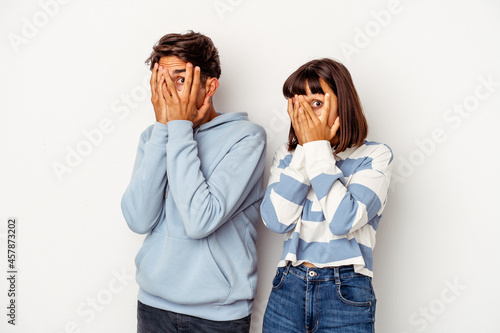 Young mixed race couple isolated on white background blink through fingers frightened and nervous.