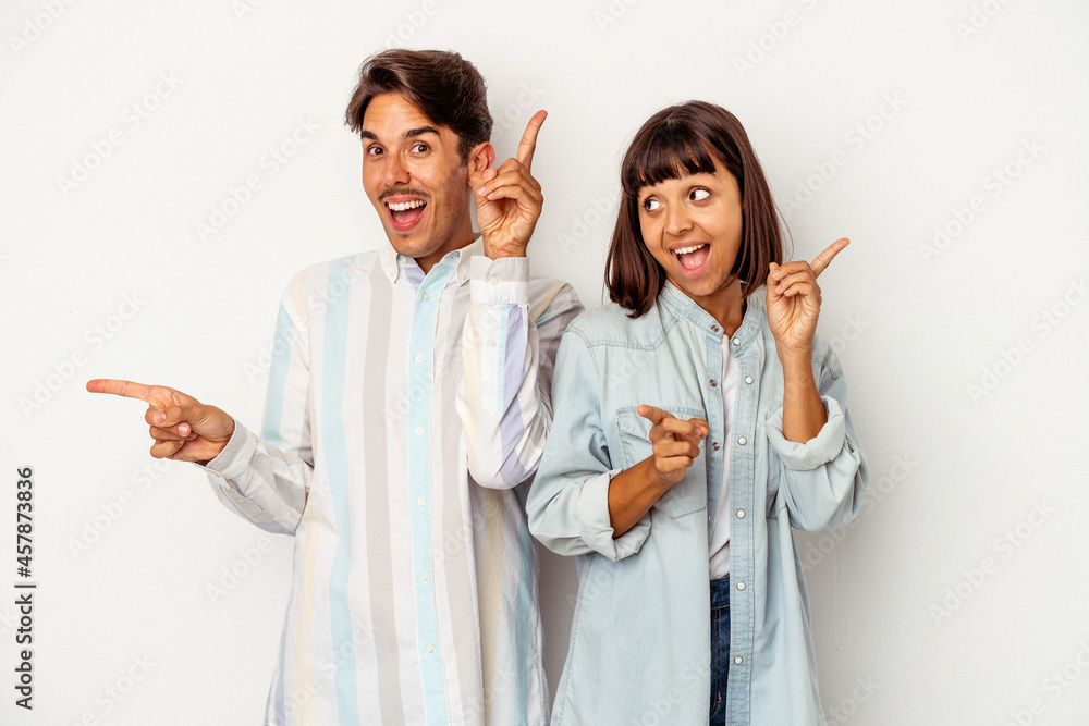 Young mixed race couple isolated on white background pointing to different copy spaces, choosing one of them, showing with finger.