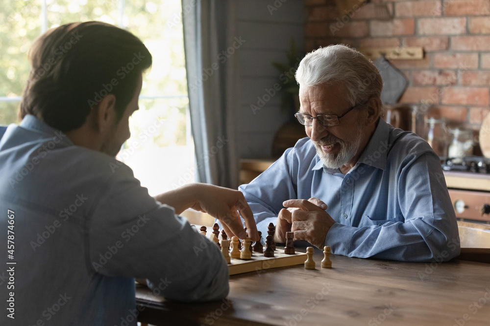 Smiling mature man in glasses with grownup son playing chess together, sitting at wooden table, senior grandfather with grandson engaged in educational activity, logic game, spending leisure time Smiling mature man in glasses with grownup son playing chess together, sitting at wooden table, senior grandfather with grandson engaged in educational activity, logic game, spending leisure time