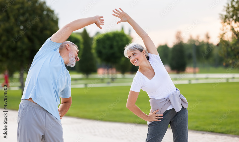 Happy smiling mature man and woman in sportswear stretching body while warming up together outdoors Happy smiling mature man and woman in sportswear stretching body while warming up together outdoors