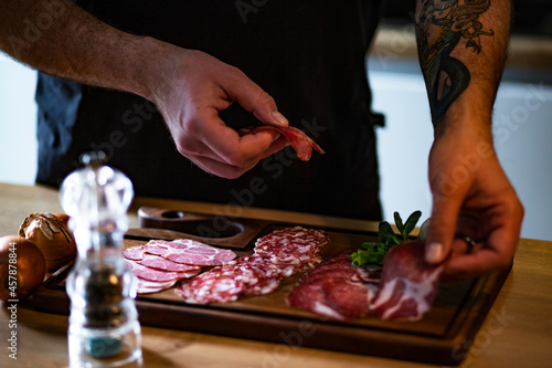 Food concept; Chef decorate ready dish with slices salami.
Young man with tattoo decorate delicious snack rustic salami with onion and cress.