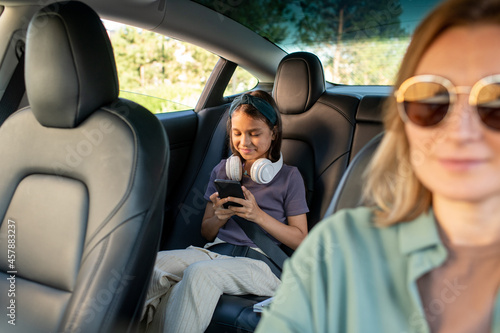 Happy schoolgirl with headphones scrolling in smartphone while sitting on backseat of car photo