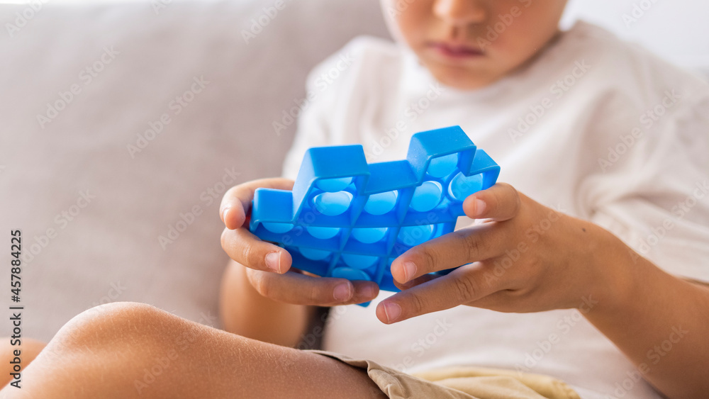 Child plays with blue pop-it in form of puzzle. Cheerful boy holds in his hands a blue toy anti-stress pop it. Little blonde boy playing with new trend sensory toy - pop it. 