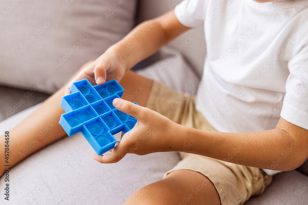 Boy lying on the couch and playing with sensory pop it fidget toy at home. Kid playes stress and anxiety relief fidgeting game. Boy with pop it a heart shape sensory toy.
