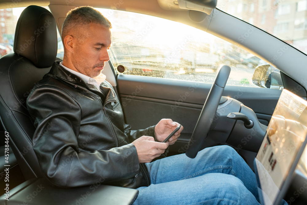 Contemporary man in blue jeans and black leather jacket texting in smartphone while sitting in electric car