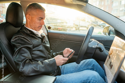 Contemporary man in blue jeans and black leather jacket texting in smartphone while sitting in electric car © pressmaster