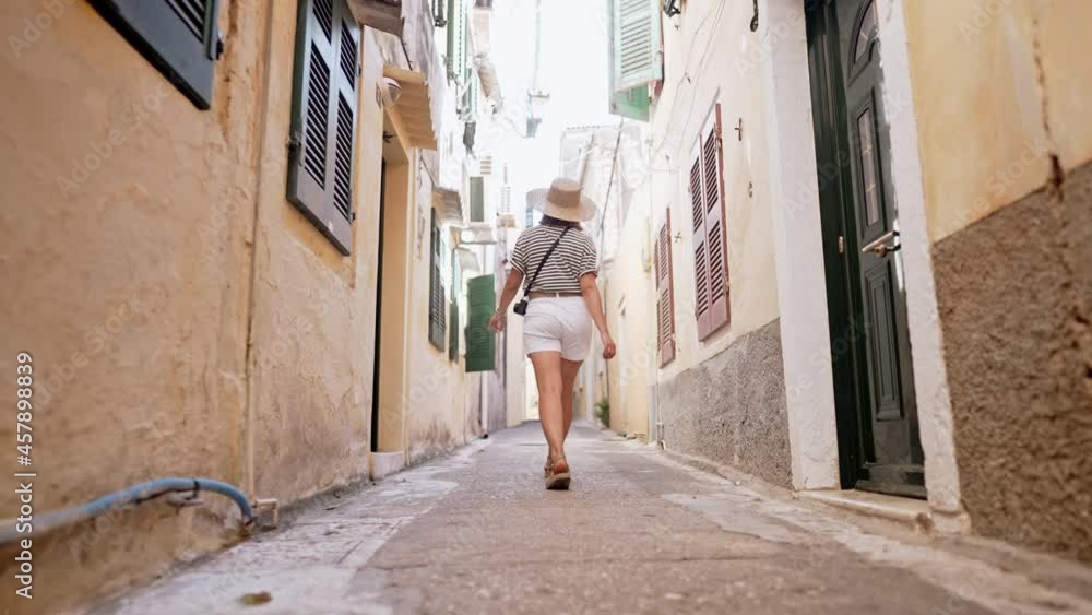 Back view of tourist woman walking alone, exploring old european city narrow streets on Greece island. Lady sightseeing local architecture. She is having good time wandering around town in summer Back view of tourist woman walking alone, exploring old european city narrow streets on Greece island. Lady sightseeing local architecture. She is having good time wandering around town in summer