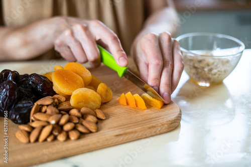 female hands preparing a healthy breakfast: cut dried fruits and nuts with a green knife: dried apricots, almonds and dates on a wooden cutting board into a glass bowl