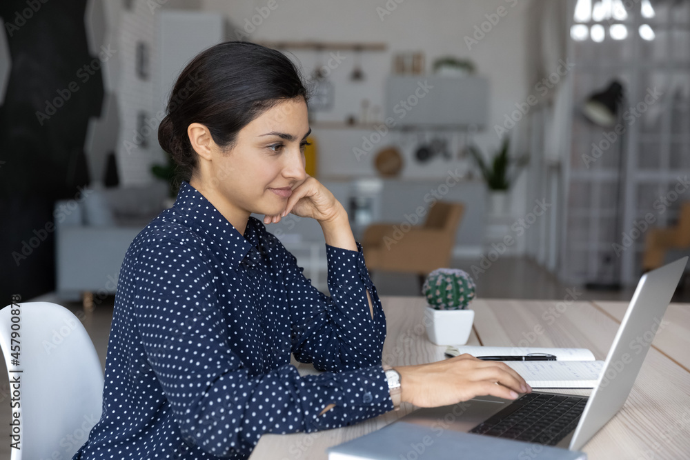 Side view confident Indian young businesswoman working on laptop online, looking at computer screen, focused woman freelancer or student typing email writing message, sitting at home office desk Side view confident Indian young businesswoman working on laptop online, looking at computer screen, focused woman freelancer or student typing email writing message, sitting at home office desk
