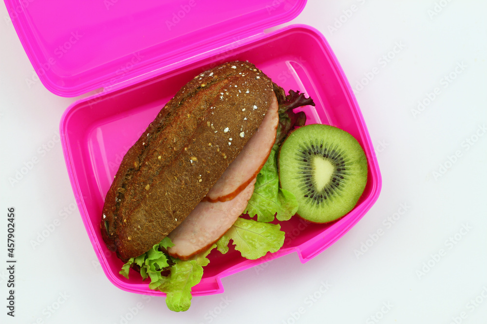 Healthy lunch box containing brown cheese sandwich with smoked pork sausage and green lettuce with kiwi fruit on white background shot from the top