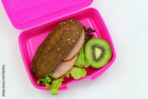 Obraz na płótnie Healthy lunch box containing brown cheese sandwich with smoked pork sausage and green lettuce with kiwi fruit on white background shot from the top
w salonie