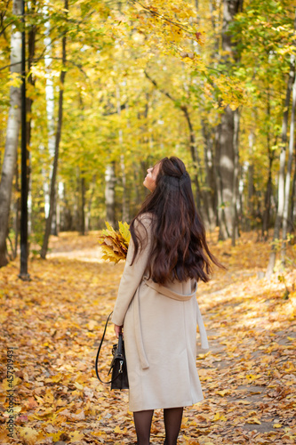 Brunette woman in autumn park and autumn leaves. Small black bag. Autumn mood. Yellow, red and green leaves. Beige coat.