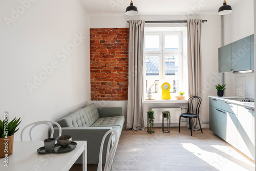 Small interior of loft apartment in industrail style with window and bricky wall. Cozy living room and kitchen with furniture, sofa and table with chairs.