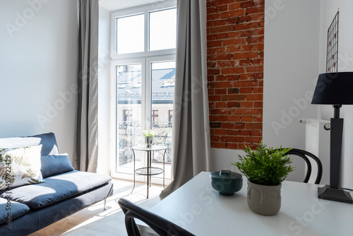 Fototapeta Naklejka Na Ścianę i Meble -  Modern interior of living room in loft apartment in industrial style with window, bricky wall and comfortable sofa. Dining room with table and chairs