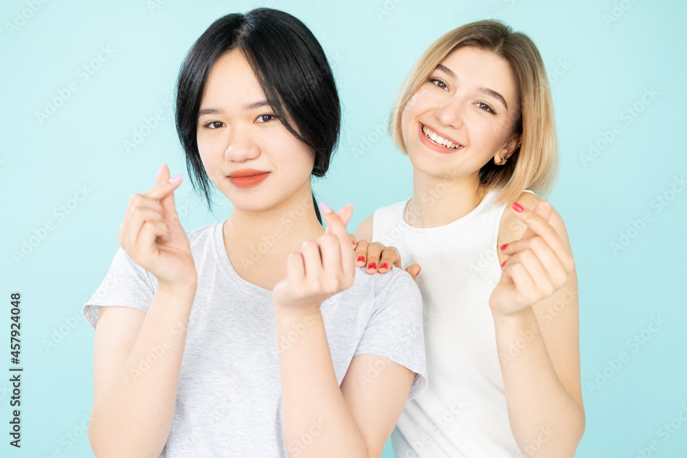 Finger heart. Happy female friends. Ethnic tolerance. International love. Pretty smiling asian caucasian women showing korean love gesture isolated blue. Finger heart. Happy female friends. Ethnic tolerance. International love. Pretty smiling asian caucasian women showing korean love gesture isolated blue.