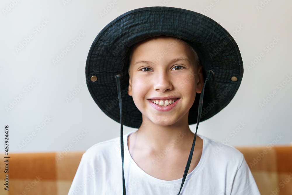 Candid shot of happy child with smiling face sitting on sofa, wearing white t-shirt and black panama hat. Portrait of a positive caucasian child sitting alone relaxing at home on weekend Candid shot of happy child with smiling face sitting on sofa, wearing white t-shirt and black panama hat. Portrait of a positive caucasian child sitting alone relaxing at home on weekend