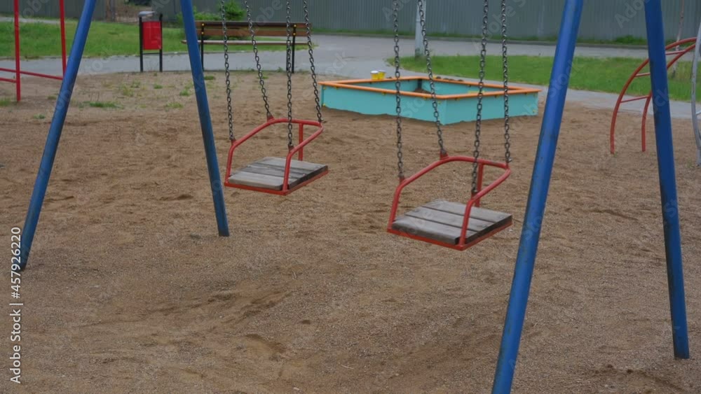 Wind sways red metal wooden swings on chains on empty children playground with sandpit ladder and bench in morning slow motion Wind sways red metal wooden swings on chains on empty children playground with sandpit ladder and bench in morning slow motion