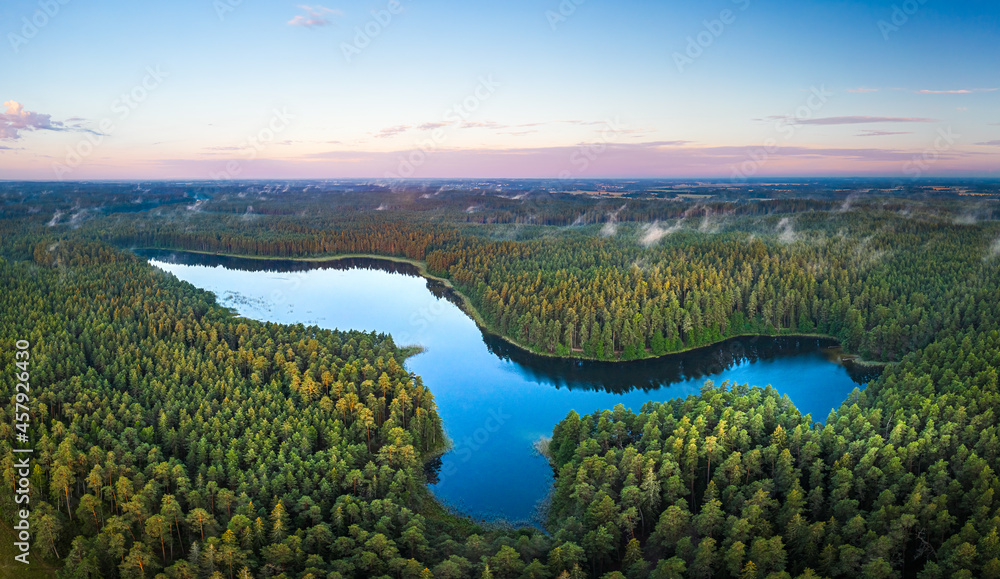 Aerial view of sunrise over a pine forest with lake. Foggy and colorful morning in countryside.