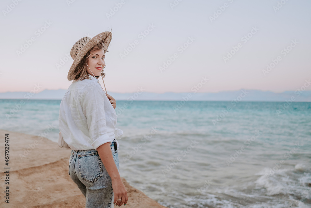 Pretty woman standing on beach near Mediterranean sea. Lady tourist in straw hat watching beautiful blue water surface, nature background. Windy weather, golden hour.