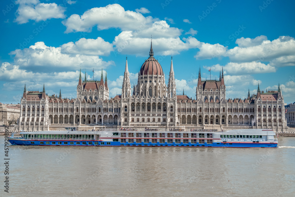 Summer view of the Hungarian Parliament built in Neo-Gothic style along the Danube river with perfect symmetry with cloudy blue sky, river cruise tourists passing by