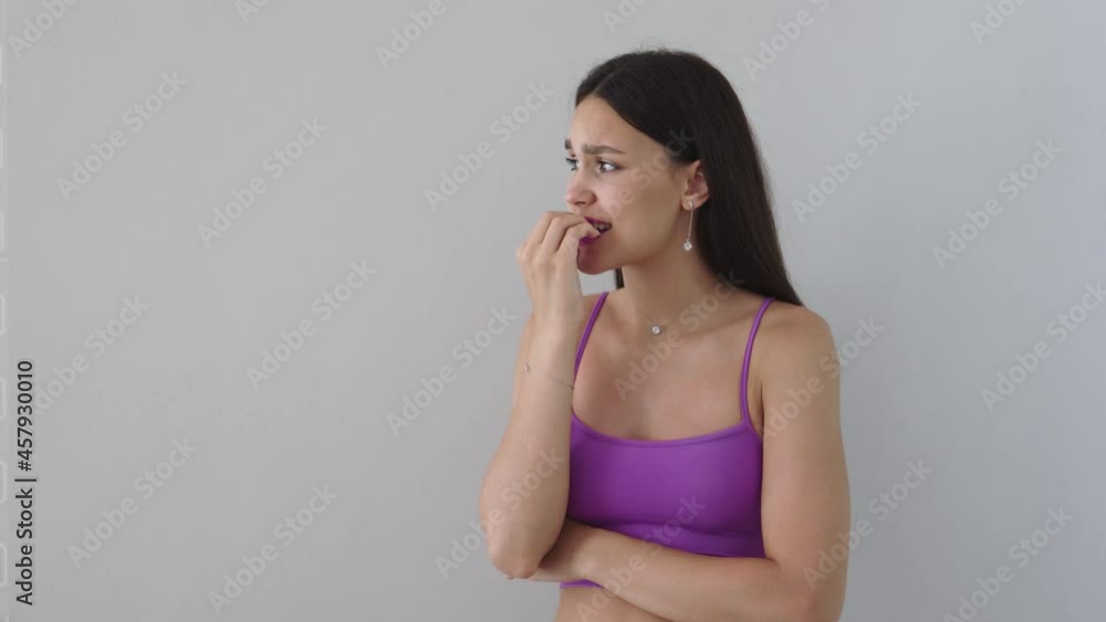 portrait of pretty slim young long brown haired caucasian female in purple clothes top and naked stomach bites long nails in mouth emotions of waiting nervous. light gray background portrait of pretty slim young long brown haired caucasian female in purple clothes top and naked stomach bites long nails in mouth emotions of waiting nervous. light gray background
