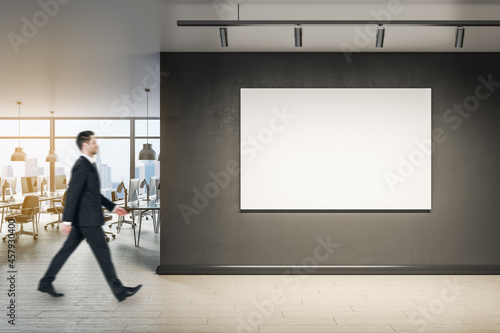 Businessman walking in modern office interior with blank wall, mock up poster and window with city view.