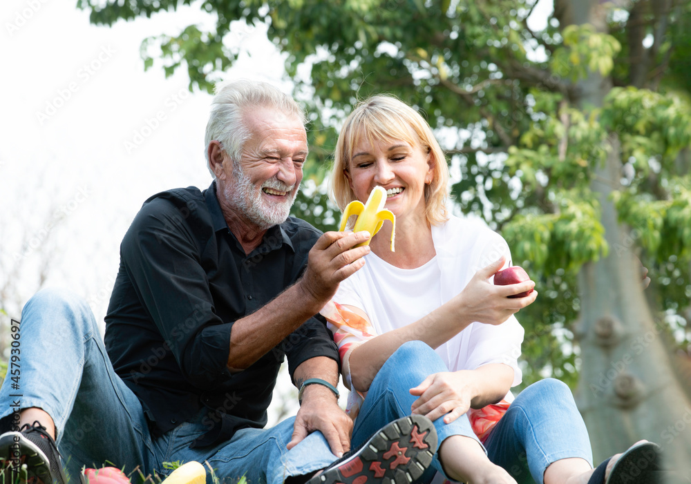Fototapeta premium Senior caucasian couple sitting on field grass and enjoy eating banana, apple together outdoors in parks. Happy mature couple eating fruits in a park. Healthy and lifestyle retired couple outdoors.
