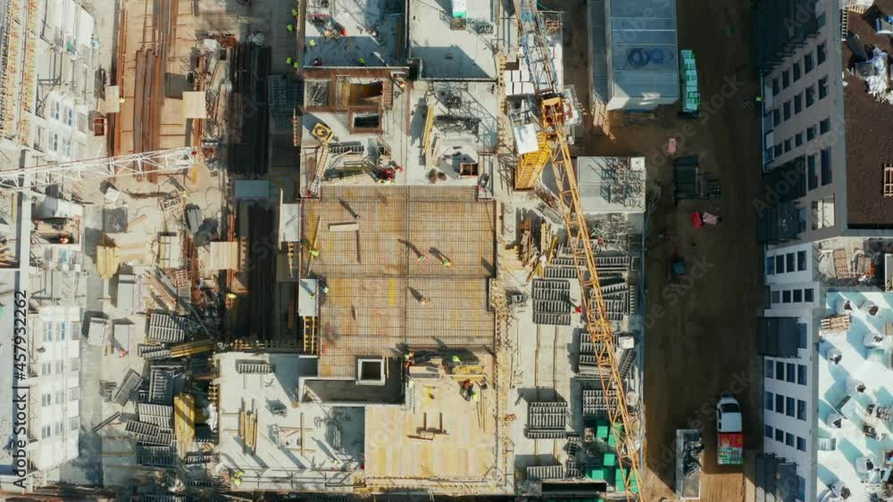 An aerial view from above down on the work of commercial and residential construction sites with tower cranes and construction workers in helmets and heavy equipment building real estate in a modern 