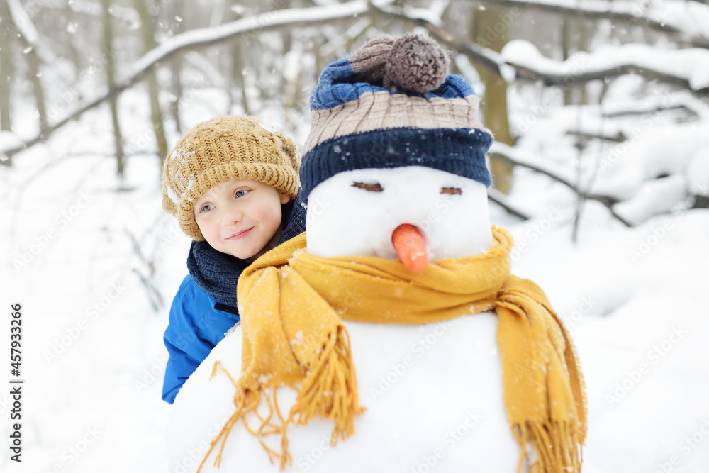 Little boy building snowman in snowy park. Child embracing snowman wearing hat and scarf. Active outdoors leisure with children in winter. Kid during stroll in a snowy winter park
