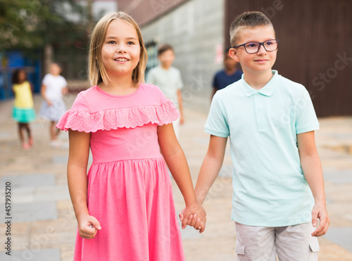 Pair of Caucasian boy and girl walking together hand in hand through city street.