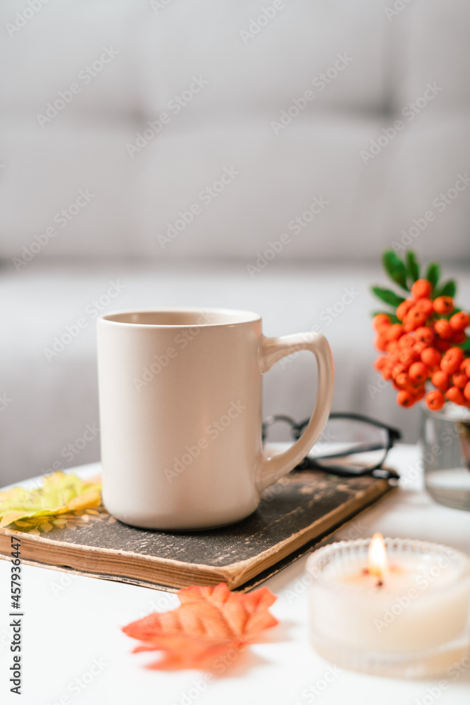 Obraz premium Still life book, candle, rowan berry and a cup of tea or coffee in the living room on a table, home decor in a cozy house. Autumn weekend concept, blanket and plaid. Fallen leaves and home decoration
