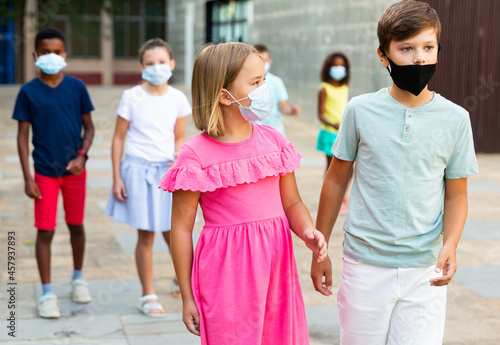 Preteen girl and boy wearing protective face masks walking along city street on summer day. Coronavirus pandemic precautions