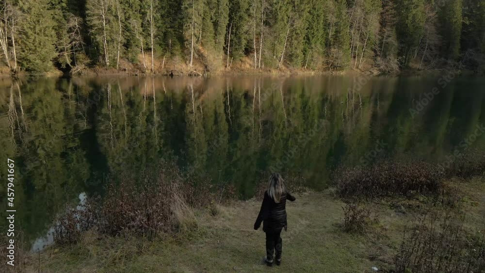 Young caucasian blonde woman dancing in front of a green lake with a forest in the background reflecting into the water, wide shot, travel concept Young caucasian blonde woman dancing in front of a green lake with a forest in the background reflecting into the water, wide shot, travel concept