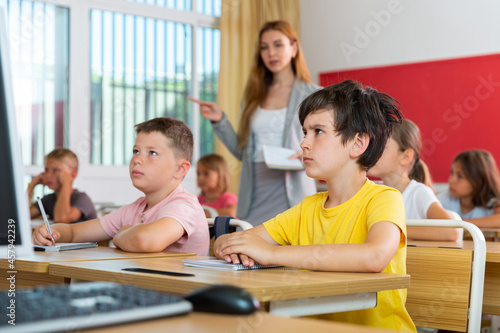 Schoolchildren sitting at desks in classroom. Female teacher staning beside desks.