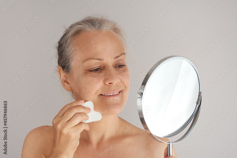 Smiling senior woman massaging her face with jade board while looking in mirror Smiling senior woman massaging her face with jade board while looking in mirror