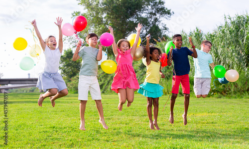 Group of happy tweenagers of different nationalities with colorful toy balloons in hands having fun together outdoors, jumping on green lawn in summertime